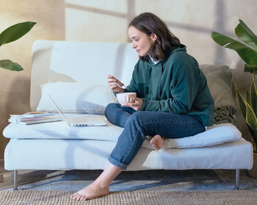 Young woman eating a bowl of ice cream on a sofa while looking at a laptop